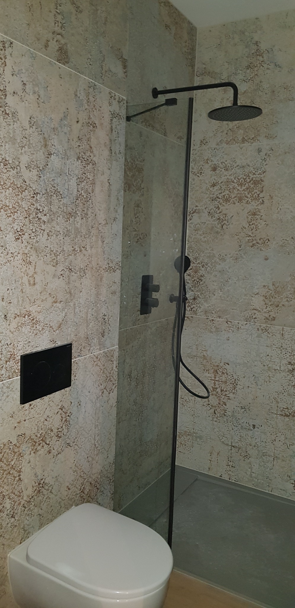 Nowoczesna kabina prysznicowa z czarną armaturą i deszczownicą, obok podwieszana toaleta, ściany wyłożone płytkami imitującymi kamień.