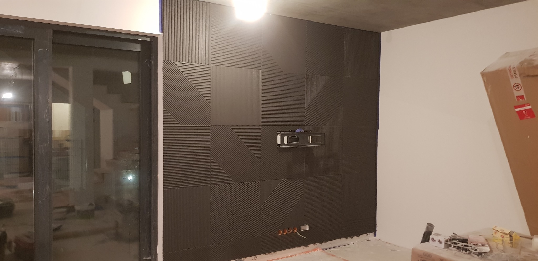 Montaż dekoracyjnych paneli ściennych z ukrytym uchwytem TV i przygotowanymi gniazdkami elektrycznymi, widok na surowy betonowy sufit i okno przesuwne