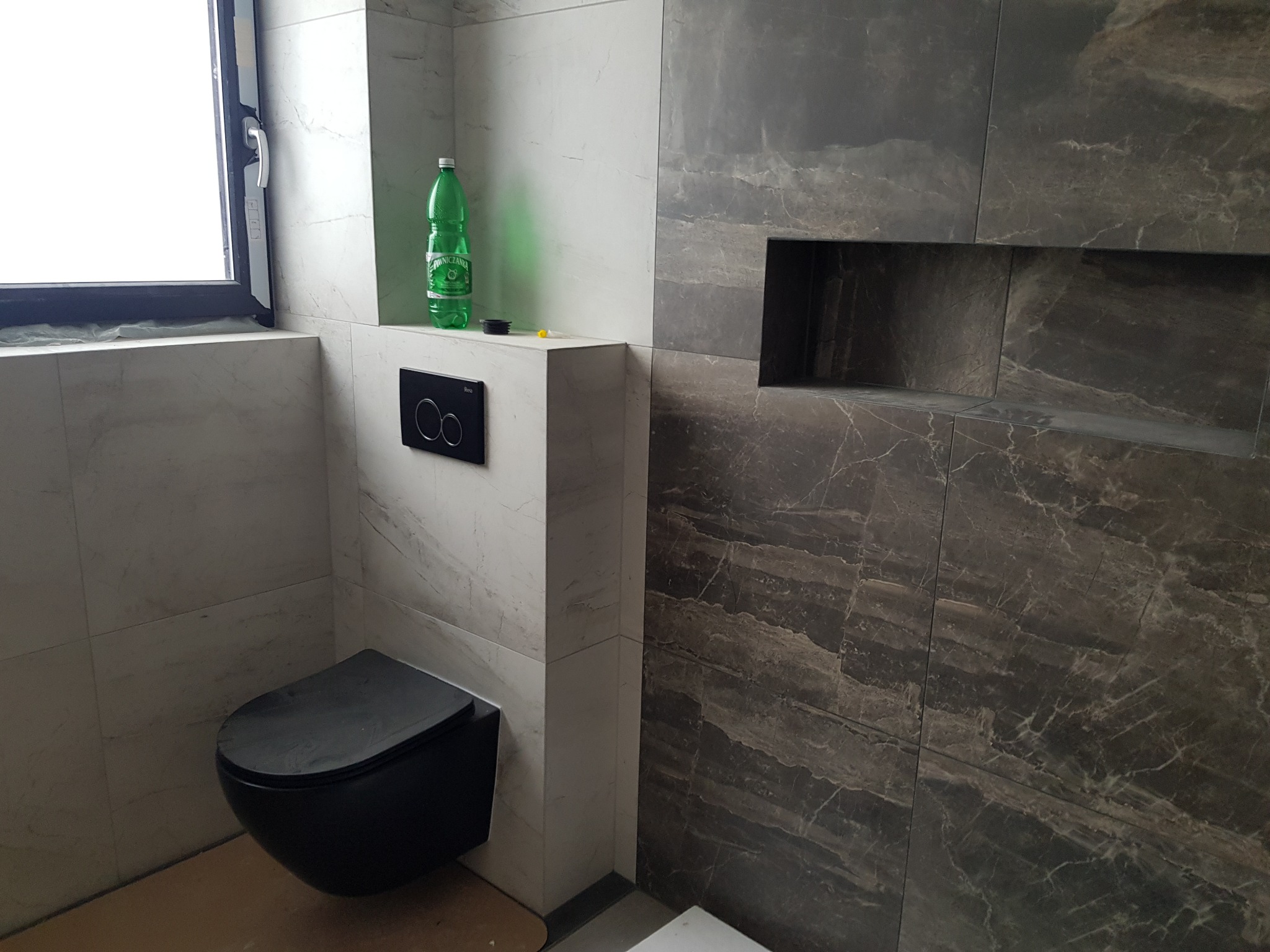 Nowoczesna łazienka z czarną, podwieszaną toaletą, wykończona płytkami imitującymi marmur w odcieniach szarości i bieli, z wnęką w ścianie i butelką wody mineralnej na zabudowie.