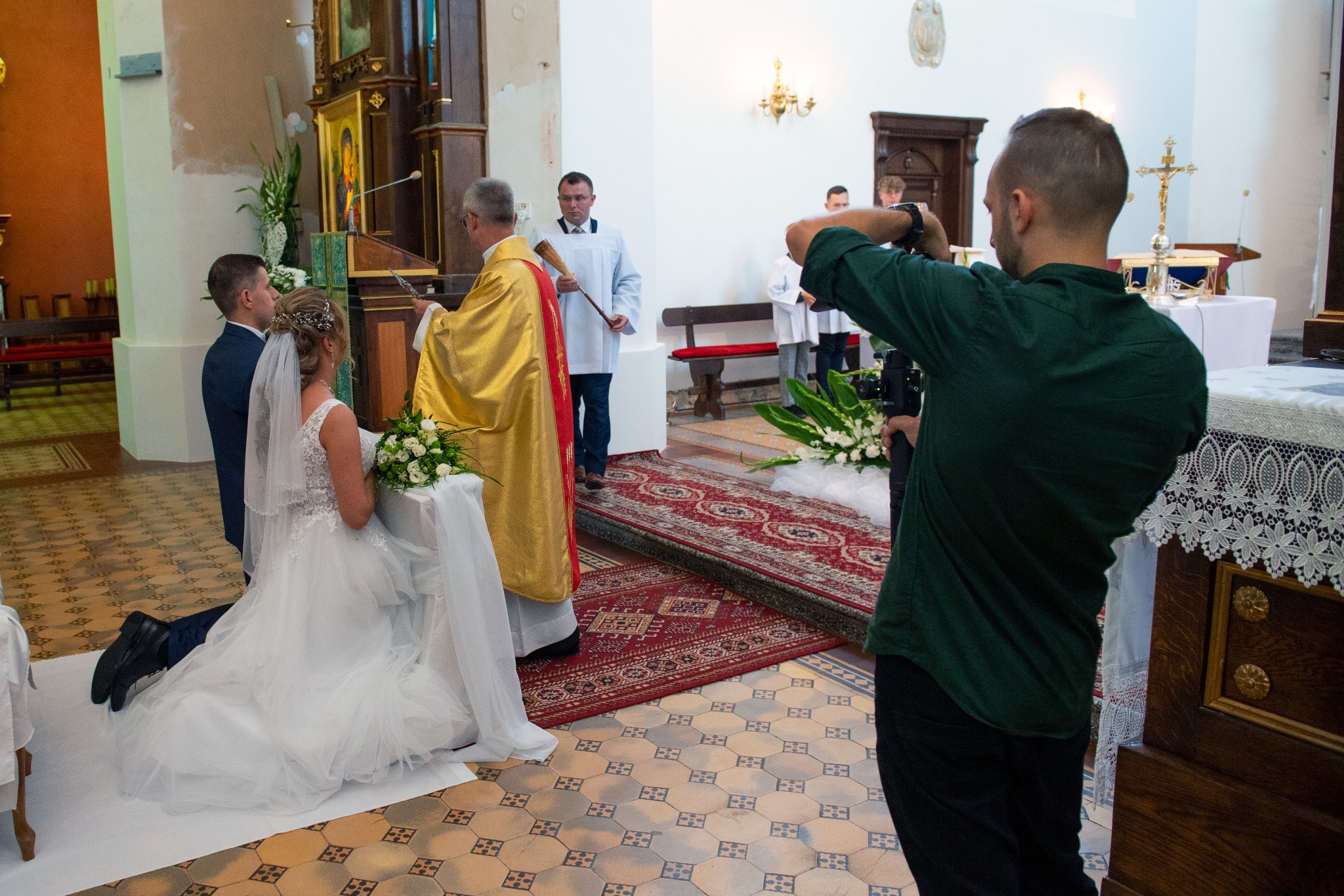 Kamerzysta filmuje ceremonię ślubną w kościele, skupiając się na klęczącej parze młodej i księdzu w złotej szacie, z widocznym dywanem w orientalne wzory.