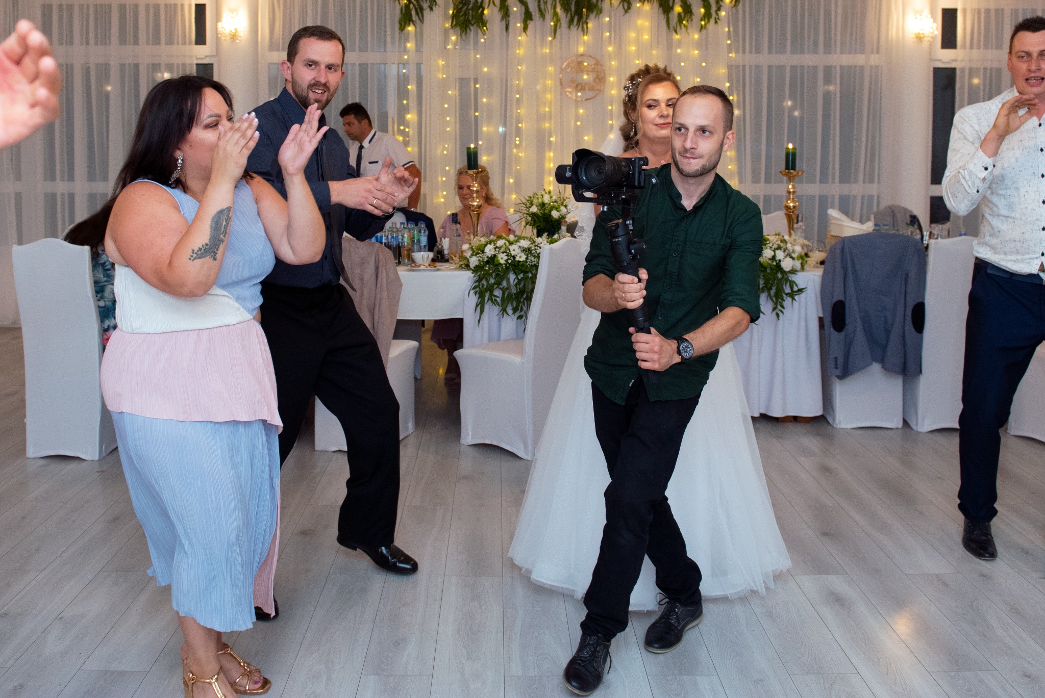 Operator kamery z gimbalem filmuje parę młodą i gości weselnych podczas zabawy na sali bankietowej z dekoracjami.