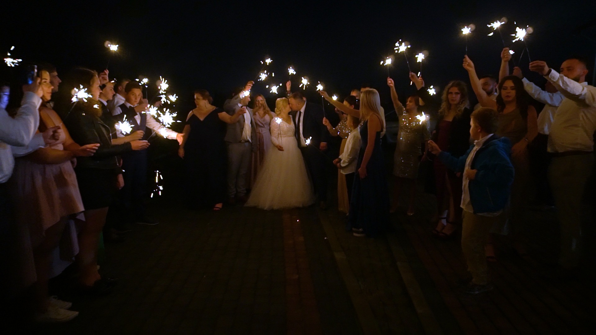 Para młoda całuje się otoczona gośćmi weselnymi trzymającymi zapalone zimne ognie w nocy.