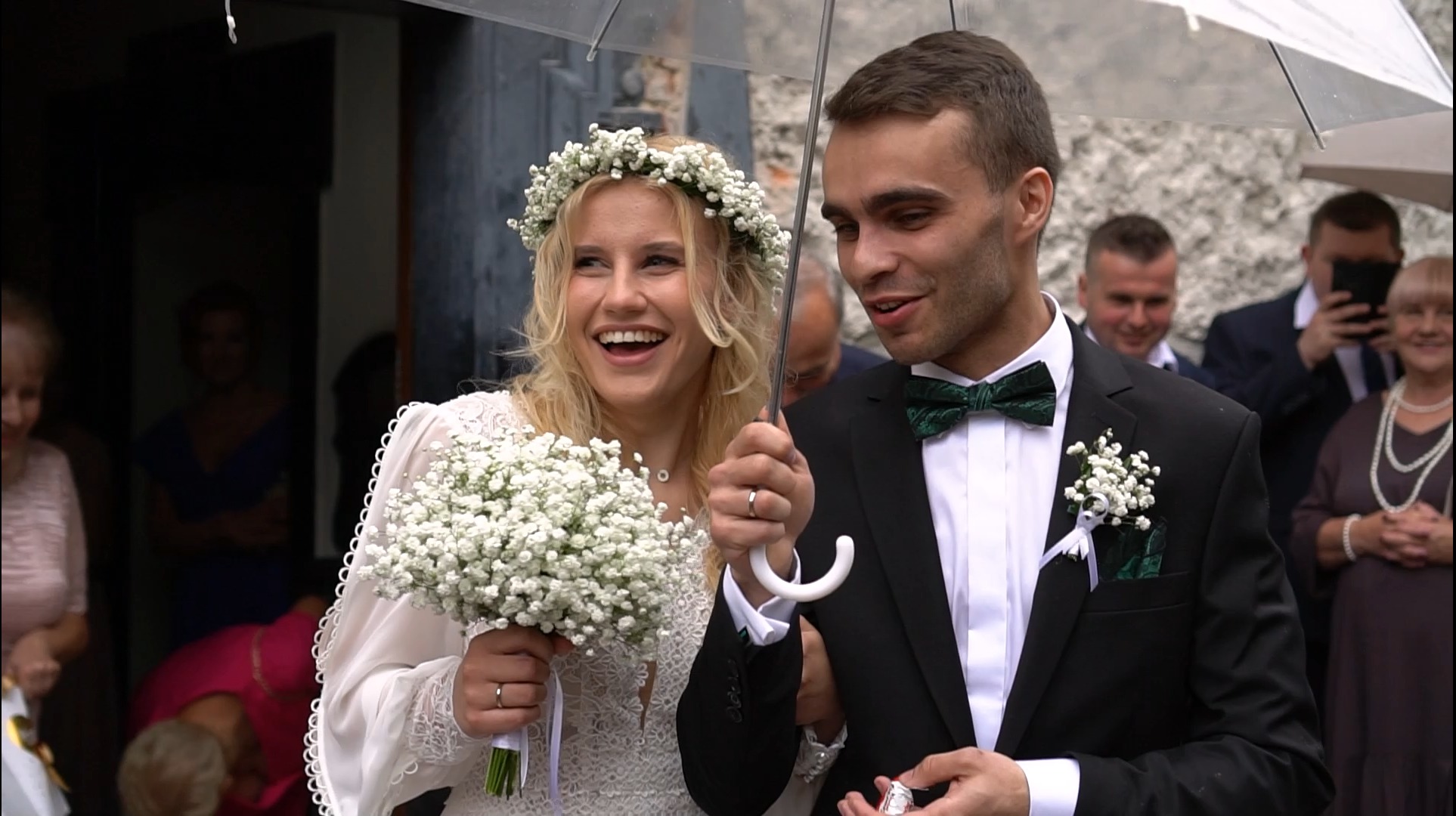 Uśmiechnięta panna młoda z bukietem gipsówki i pan młody pod przezroczystym parasolem w dniu ślubu, widoczni goście w tle.