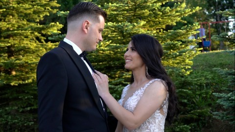 Uśmiechnięta panna młoda poprawia muchę panu młodemu na tle zielonych drzew iglastych.