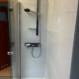 Nowoczesna kabina prysznicowa z chromowaną armaturą i szklanymi drzwiami, widok na drewniane drzwi i podłogę poza kabiną.