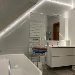 Nowoczesna łazienka na poddaszu z oświetleniem LED, białą wanną, grzejnikiem drabinkowym i szafką z umywalką, uzupełniona krzesłem barowym z ręcznikami.