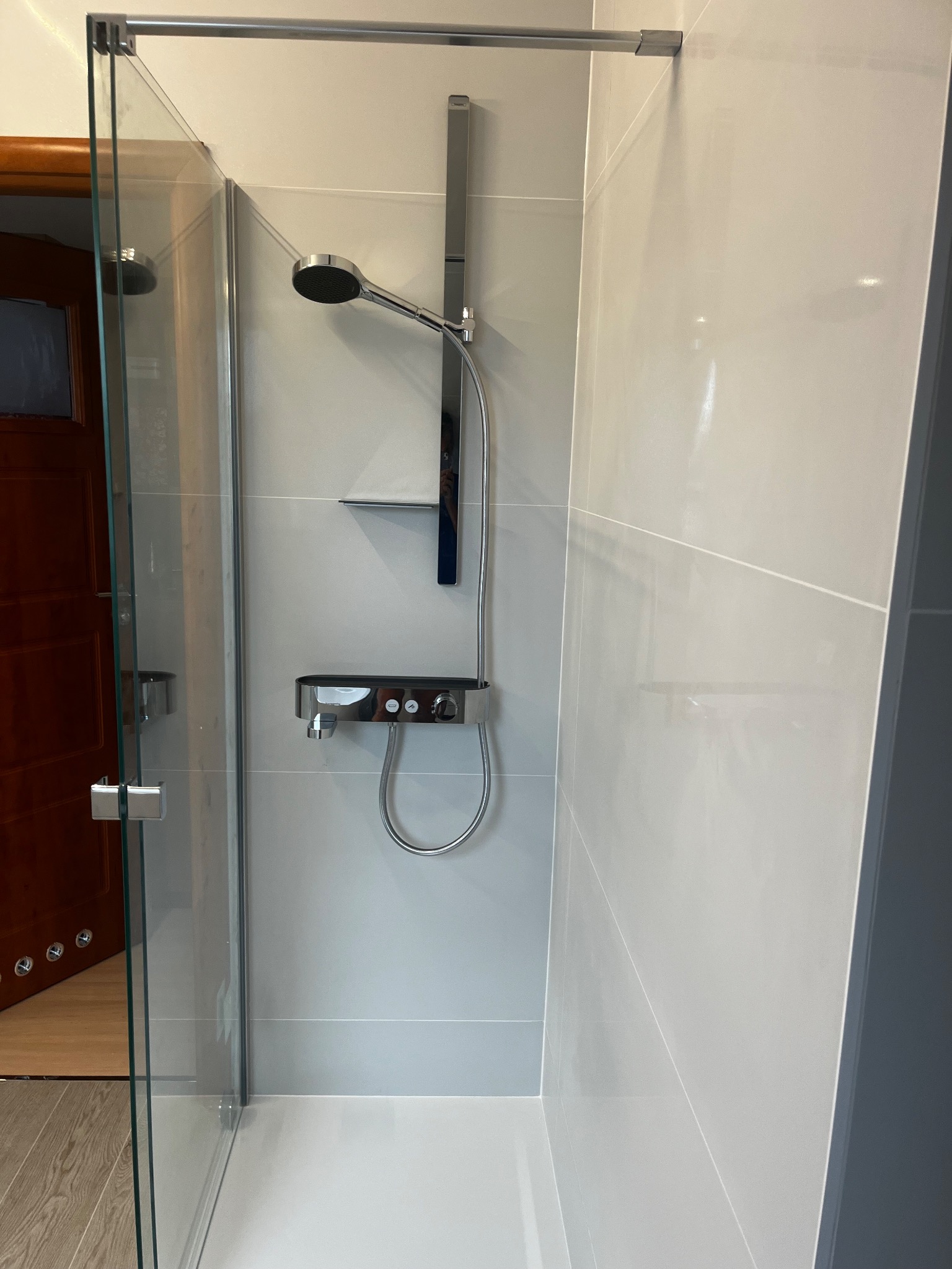 Nowoczesna kabina prysznicowa z chromowaną armaturą i szklanymi drzwiami, widok na drewniane drzwi i podłogę poza kabiną.