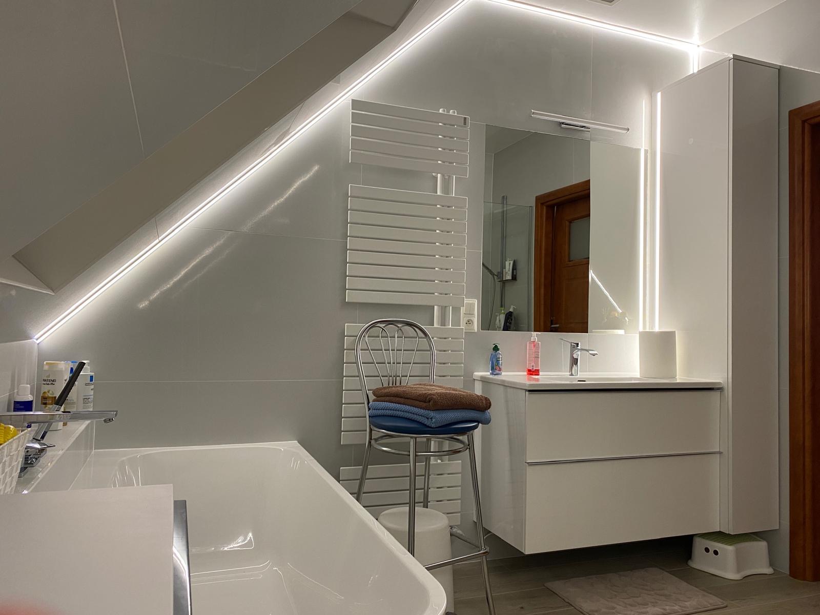 Nowoczesna łazienka na poddaszu z oświetleniem LED, białą wanną, grzejnikiem drabinkowym i szafką z umywalką, uzupełniona krzesłem barowym z ręcznikami.