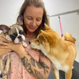 Kobieta z tatuażami trzyma buldoga francuskiego, obok stoi Shiba Inu na stole do pielęgnacji psów z metalowym ramieniem