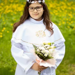 Dziewczynka w białej albie z bukietem kwiatów na tle łąki z żółtymi kwiatami, ubrana w wianek z białych kwiatów i okulary.