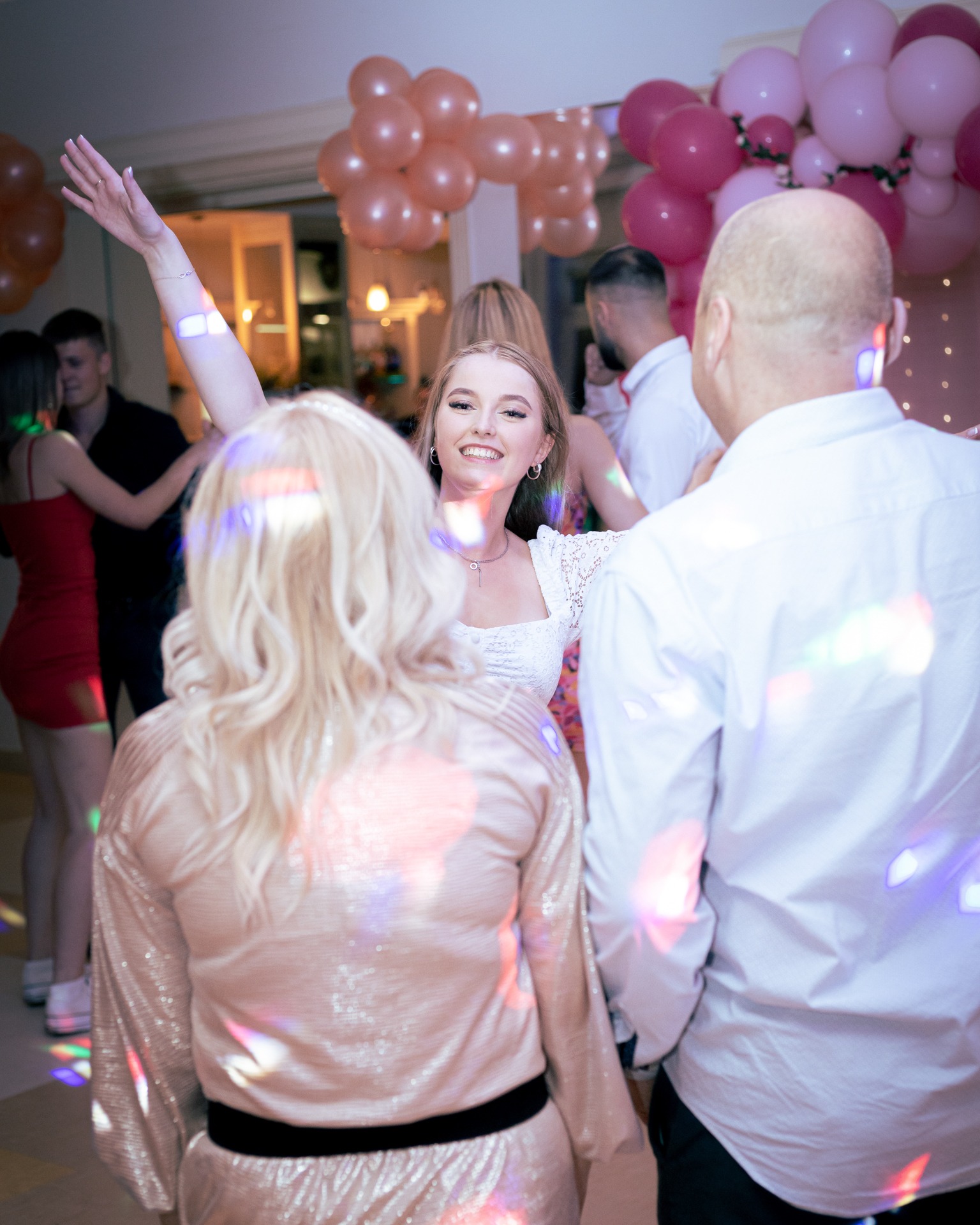 Młoda kobieta w białej sukience tańczy na imprezie, w tle dekoracja z różowych balonów i bawiący się ludzie, oświetlenie dyskotekowe tworzy kolorowe refleksy.