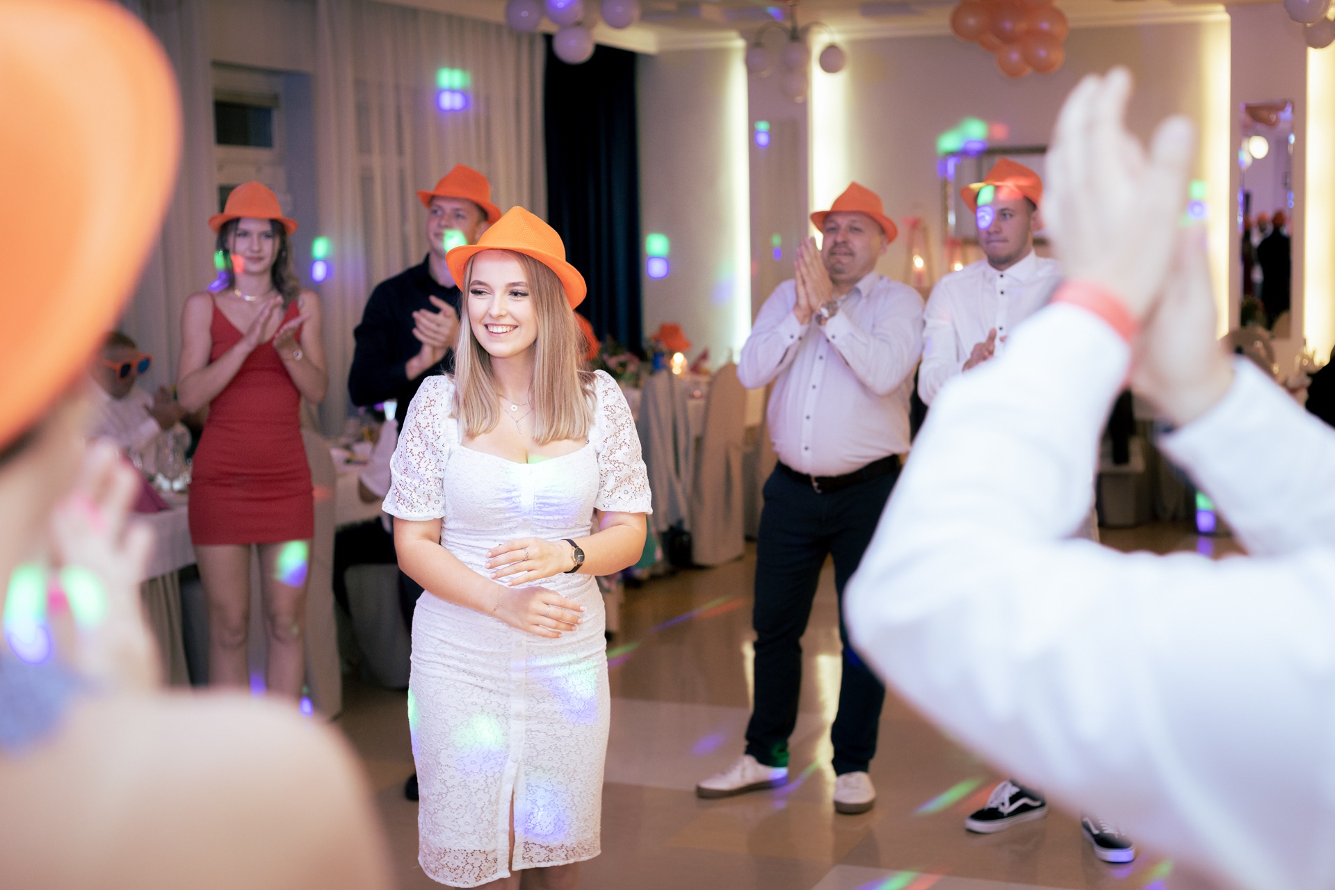 Uśmiechnięta kobieta w białej koronkowej sukience i pomarańczowym kapeluszu na imprezie, otoczona przez osoby klaszczące w dłonie, oświetlone kolorowymi światłami.