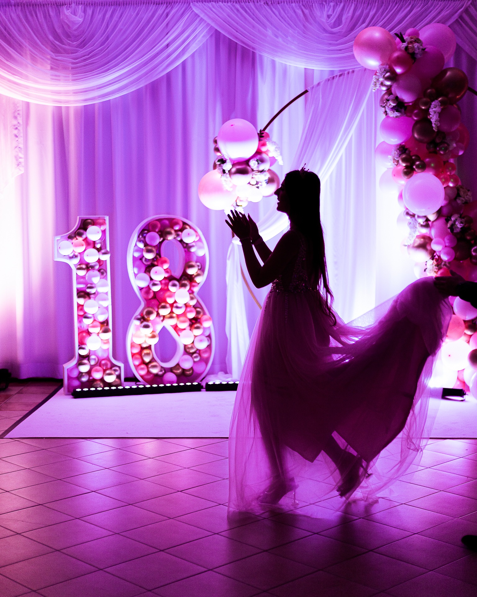 Sylwetka dziewczyny w tiulowej sukni na tle dekoracji urodzinowej z liczbą 18 wypełnioną balonami w odcieniach różu i złota, całość oświetlona fioletowym światłem.