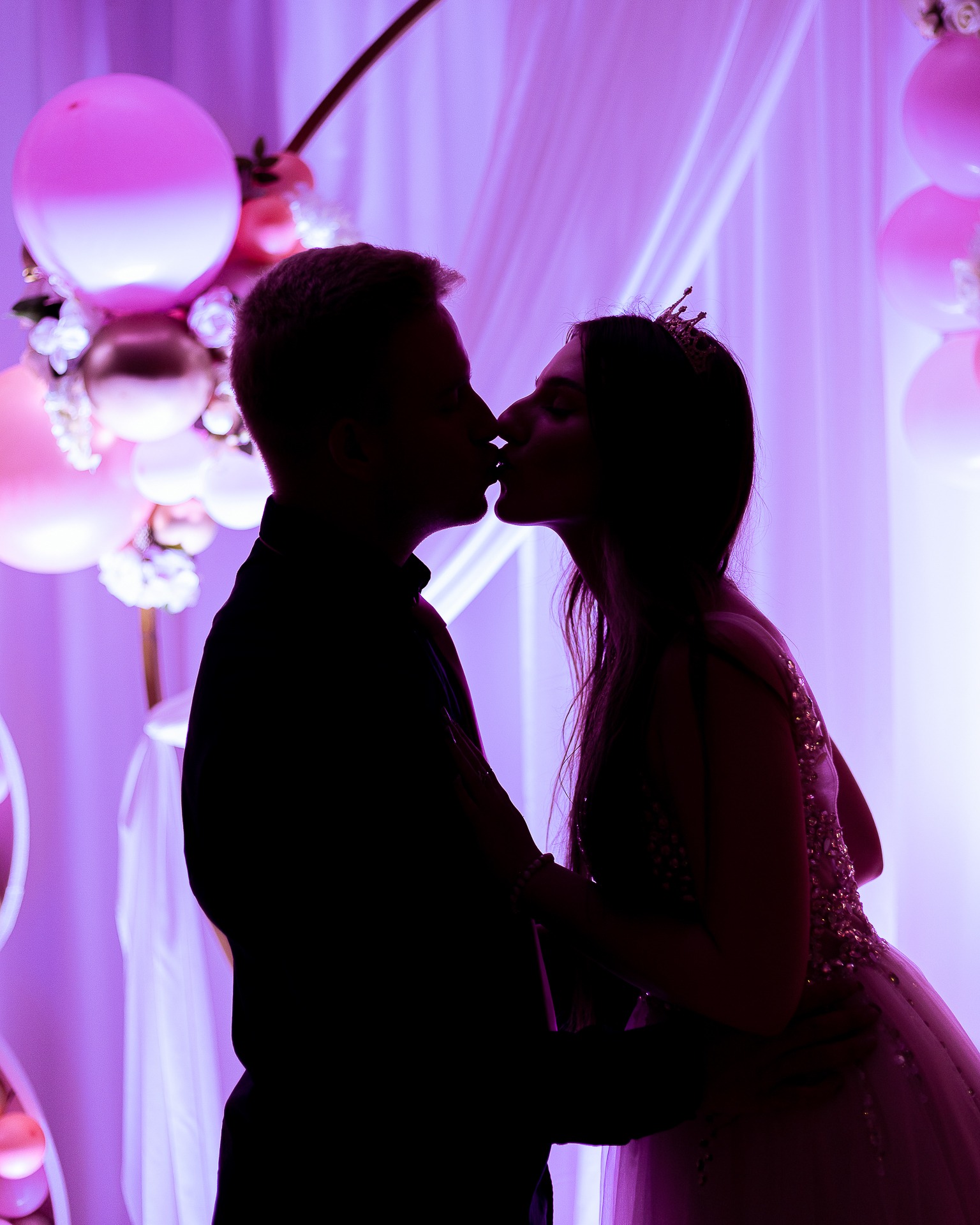 Silhouettes of a kissing couple against a backdrop of pink and white balloons and draped fabric, illuminated with purple lighting.
