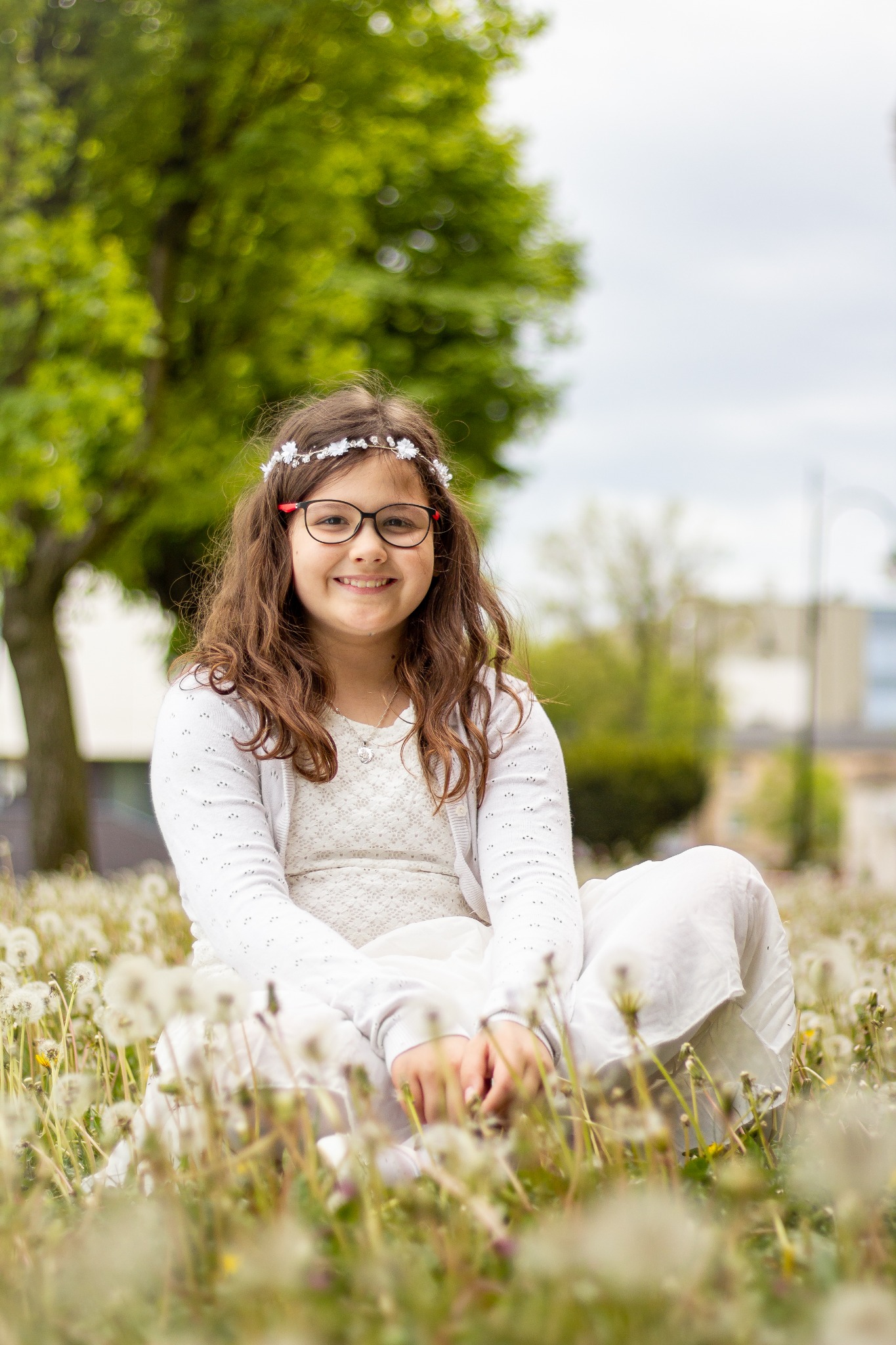 Dziewczynka w okularach i białej sukience z wiankiem na głowie siedzi na łące dmuchawców w Gnieźnie, uśmiechając się do obiektywu.