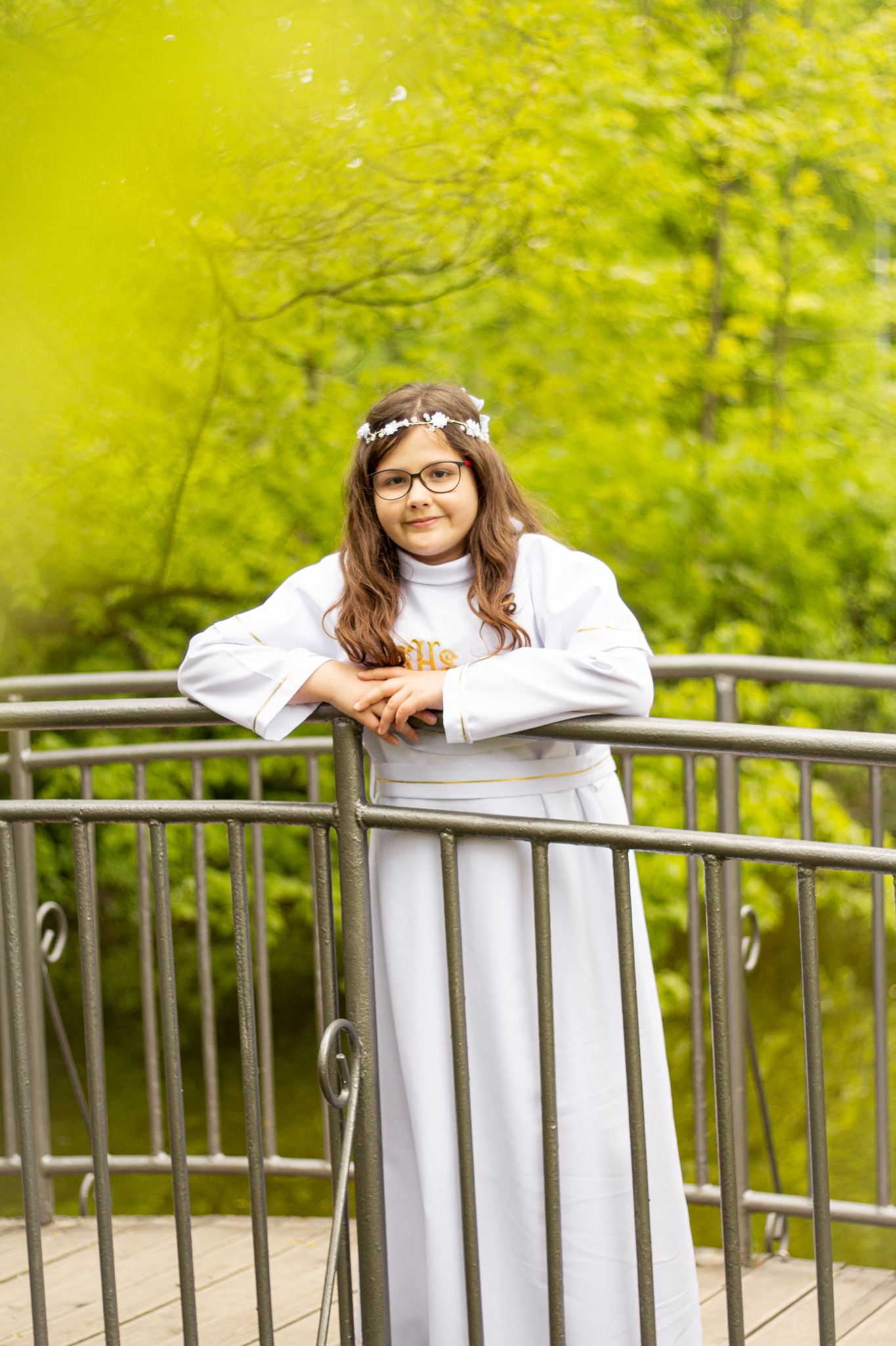 Dziewczynka w białej albie komunijnej, z wiankiem na głowie i okularach, opiera się o metalową barierkę mostku na tle zieleni.
