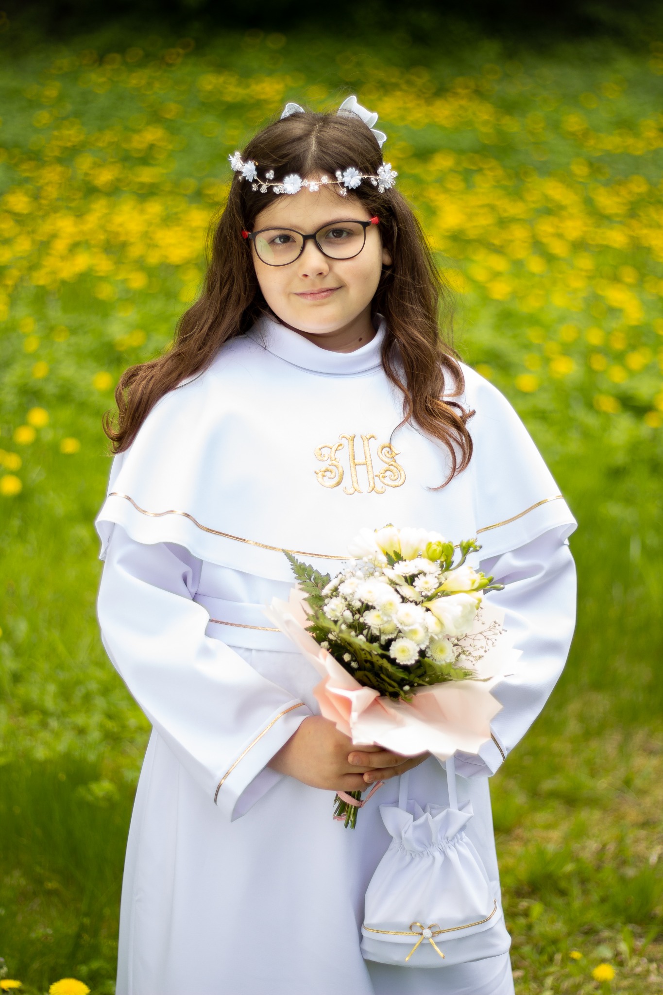 Dziewczynka w białej albie z bukietem kwiatów na tle łąki z żółtymi kwiatami, ubrana w wianek z białych kwiatów i okulary.
