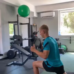 Młody mężczyzna w turkusowej koszulce i białych skarpetach wykonuje wykrok z obciążeniem w domowej siłowni; w tle widoczny sprzęt do ćwiczeń siłowych, piłka gimnastyczna i lustro.