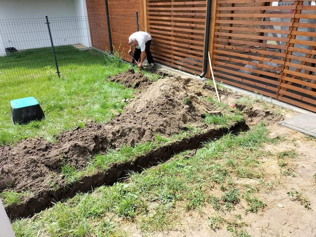 Wykopany rów w trawniku pod system nawadniania, widoczny mężczyzna kopiący łopatą, drewniany płot w tle.