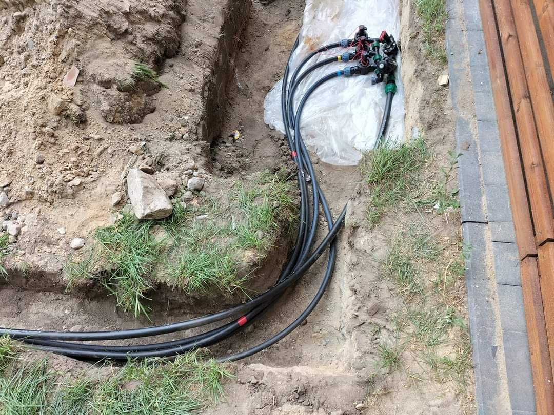 Wykonany wykop pod system nawadniania z widocznymi rurami i rozdzielaczem na folii, tuż przy drewnianym ogrodzeniu i kostce brukowej.