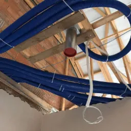 Niebieskie rury wentylacyjne przymocowane do drewnianej konstrukcji dachu z widocznym fragmentem ściany z cegieł i surowego tynku, z metalowym wylotem wentylacyjnym.