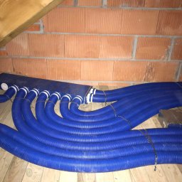 Eko-Home Tomasz Łuczak - Niebieskie, karbowane rury wentylacyjne ułożone równolegle na poddaszu, widoczne elementy mocujące i fragment ściany z czerwonej cegły.