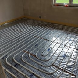 Eko-Home Tomasz Łuczak - Białe rury układane w pętle na folii z siatką, przygotowane do zalania wylewką w surowym wnętrzu z oknem.