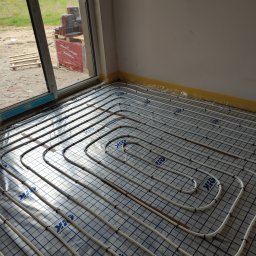 Eko-Home Tomasz Łuczak - Białe rury systemu rozprowadzone na folii z niebieską siatką w nowym budynku, widoczne płytki na palecie za oknem.