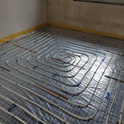 Eko-Home Tomasz Łuczak - Rozłożona instalacja ogrzewania podłogowego z białych rurek na folii z niebieską kratką, przymocowana czarnymi klipsami, widoczne brązowe listwy montażowe, żółta folia zabezpieczająca przy ścianach...