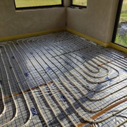 Eko-Home Tomasz Łuczak - Białe rurki systemu grzewczego ułożone w pętle na folii z niebieską kratką, widoczne okna i surowe ściany w tle.