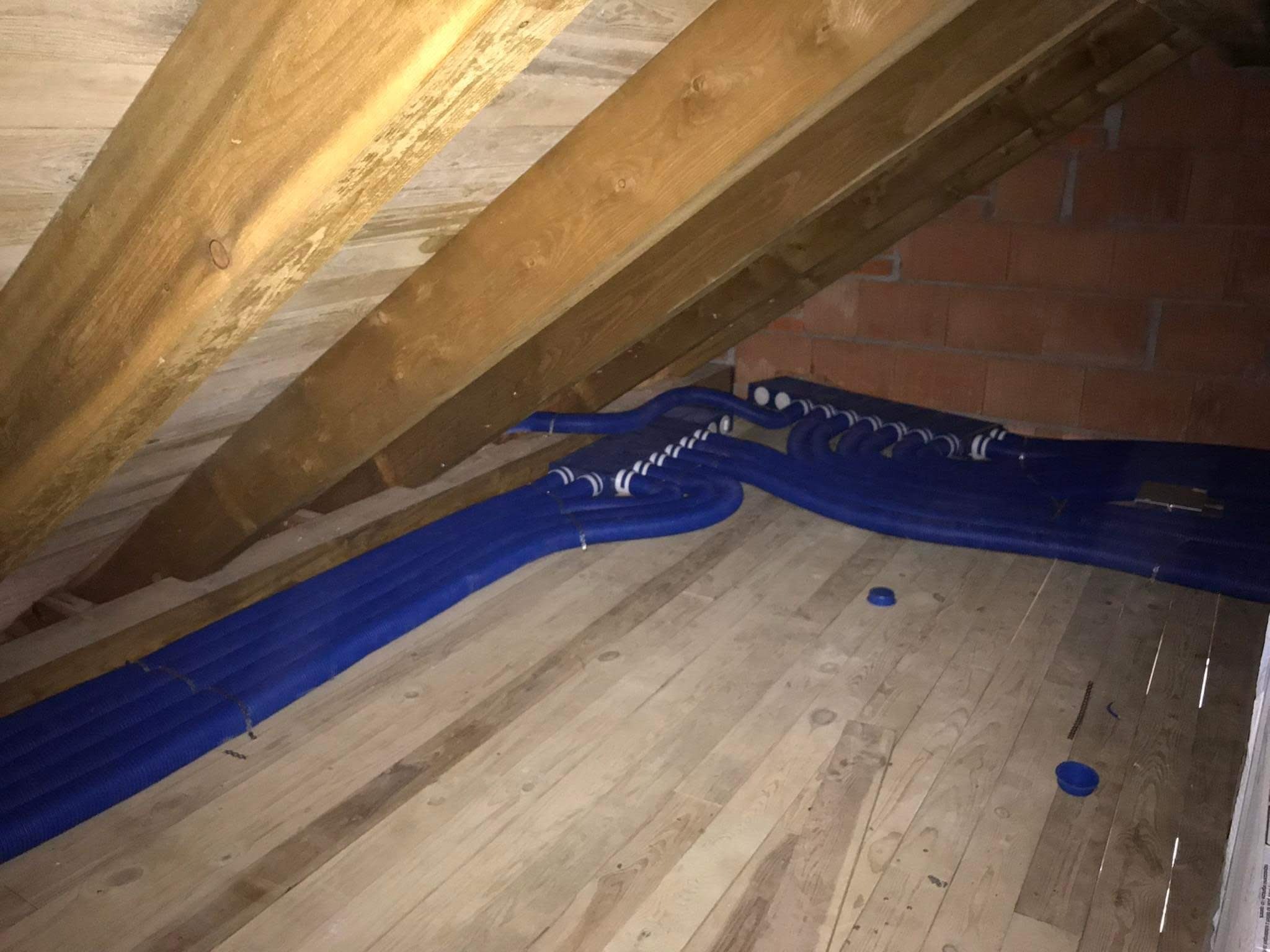 Niebieskie, karbowane rury wentylacyjne ułożone na drewnianej podłodze poddasza, widoczne krokwie i fragment ściany z czerwonej cegły.