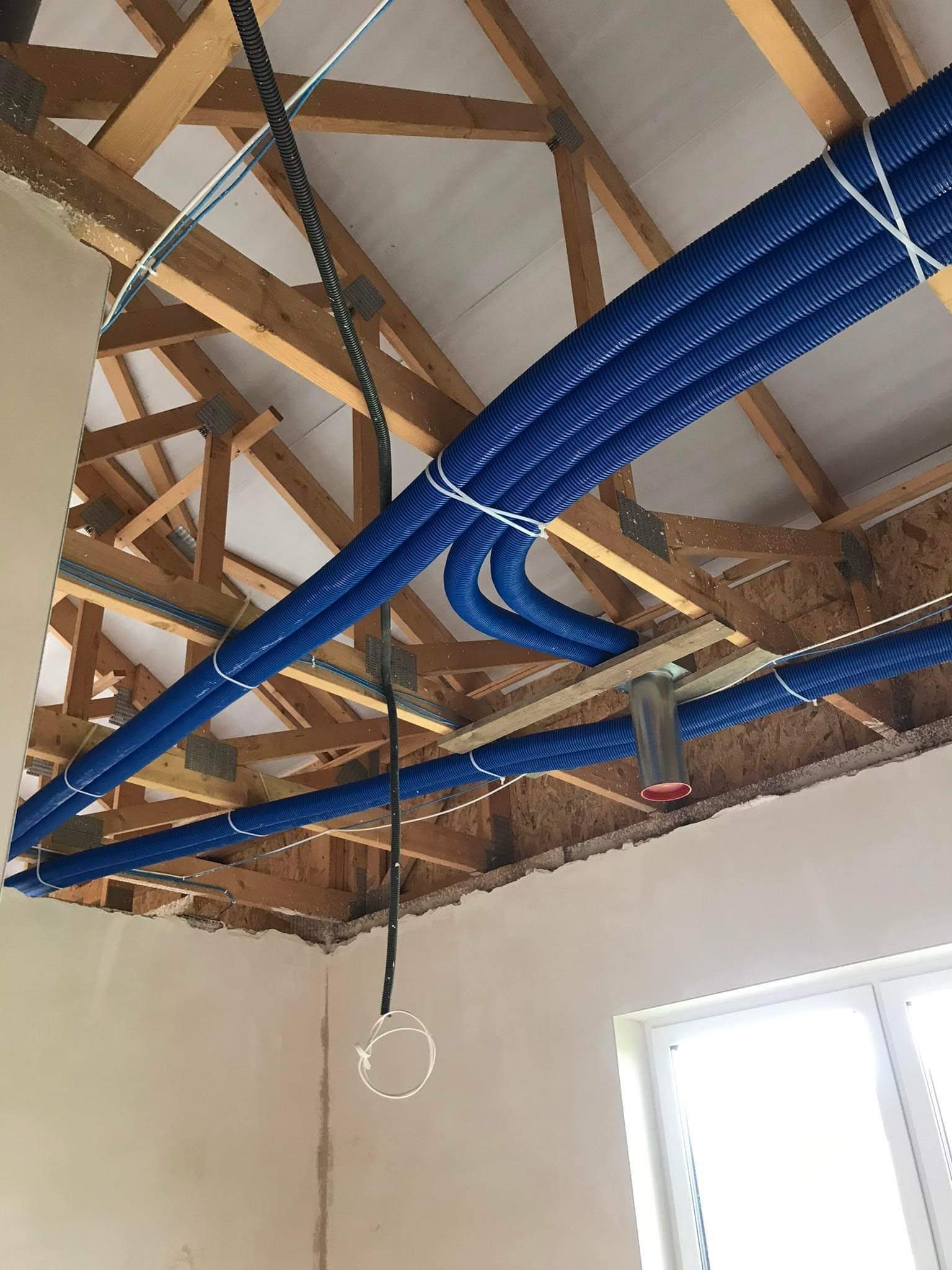 Niebieskie, karbowane rury wentylacyjne zamontowane pod drewnianą konstrukcją dachu, zabezpieczone białymi opaskami zaciskowymi, widoczne elementy instalacji i fragment surowej ściany z oknem.
