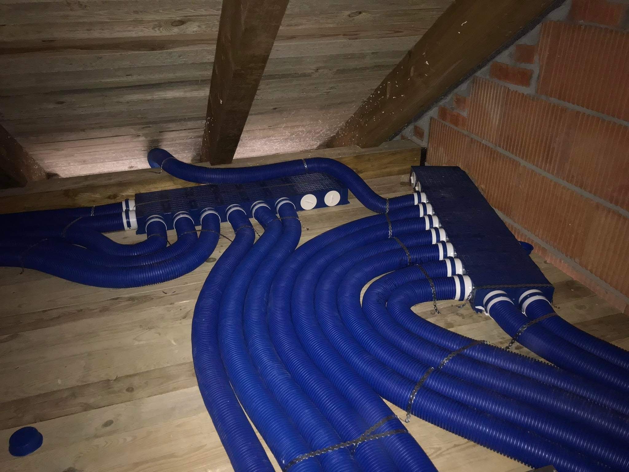 System niebieskich, karbowanych rur wentylacyjnych rozchodzących się z dwóch kolektorów na poddaszu z drewnianą podłogą i ceglaną ścianą w tle.
