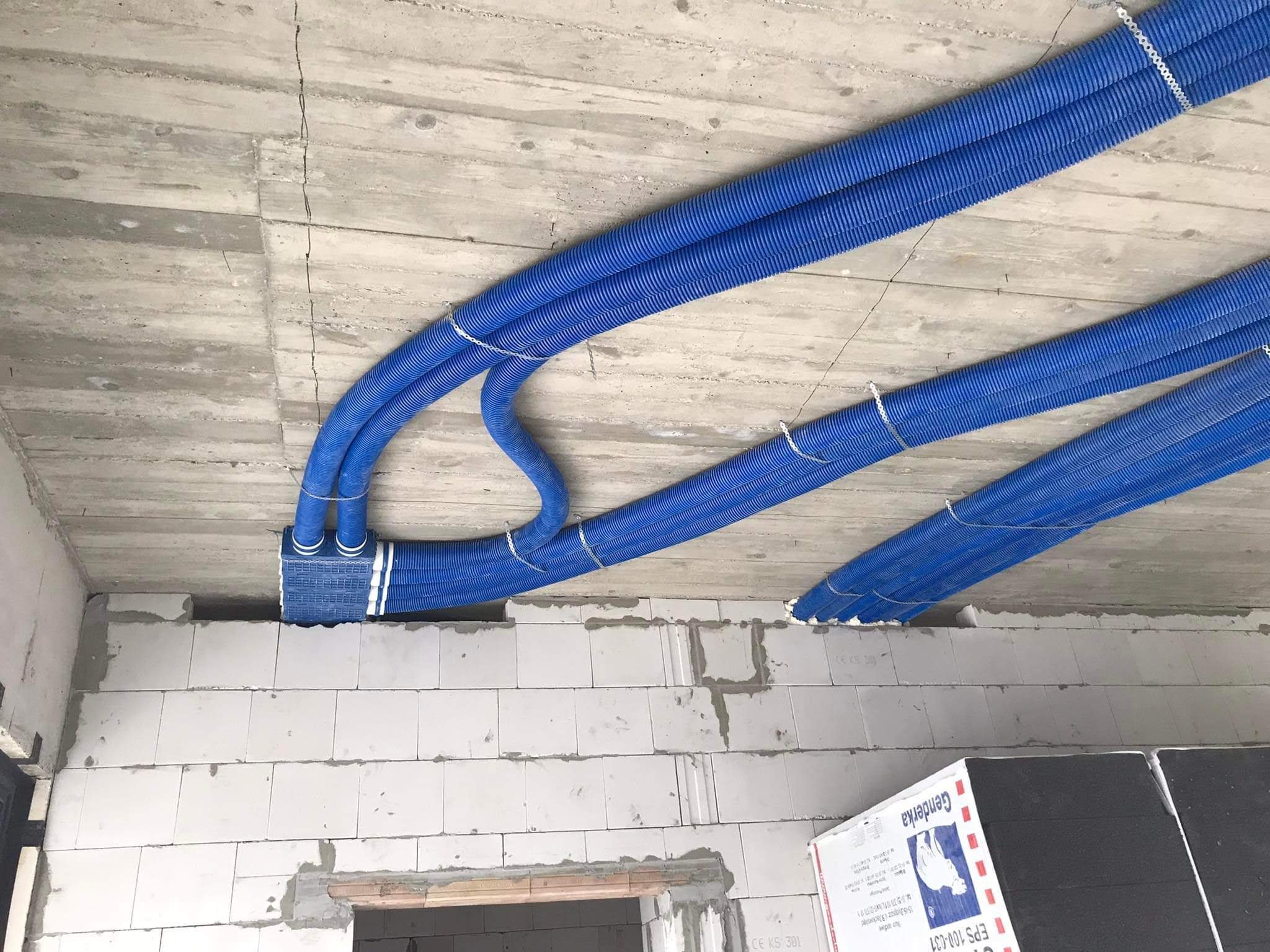 Niebieskie, karbowane rury wentylacyjne przymocowane do surowego, betonowego sufitu, częściowo ukryte w ścianie z białych bloczków.