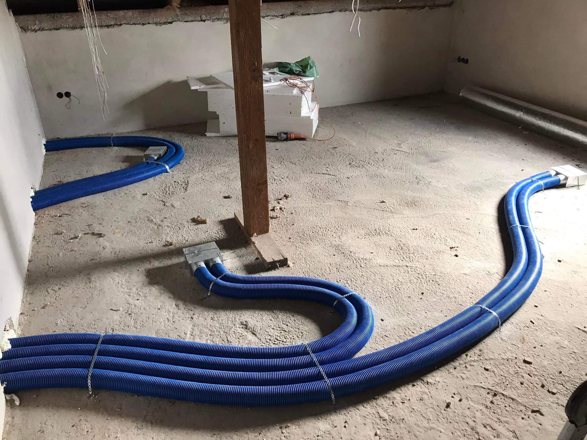 Niebieskie, karbowane rury wentylacyjne prowadzone po surowym betonowym podłożu w trakcie instalacji. Widoczne metalowe skrzynki rozdzielcze i elementy mocujące rury.