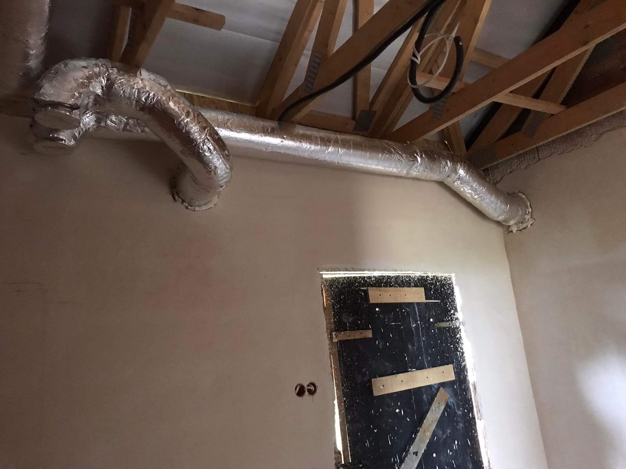 Srebrna, izolowana rura wentylacyjna zamontowana pod drewnianą konstrukcją dachu, widoczne przygotowane otwory na gniazdka elektryczne i prowizoryczne zabezpieczenie otworu drzwiowego.