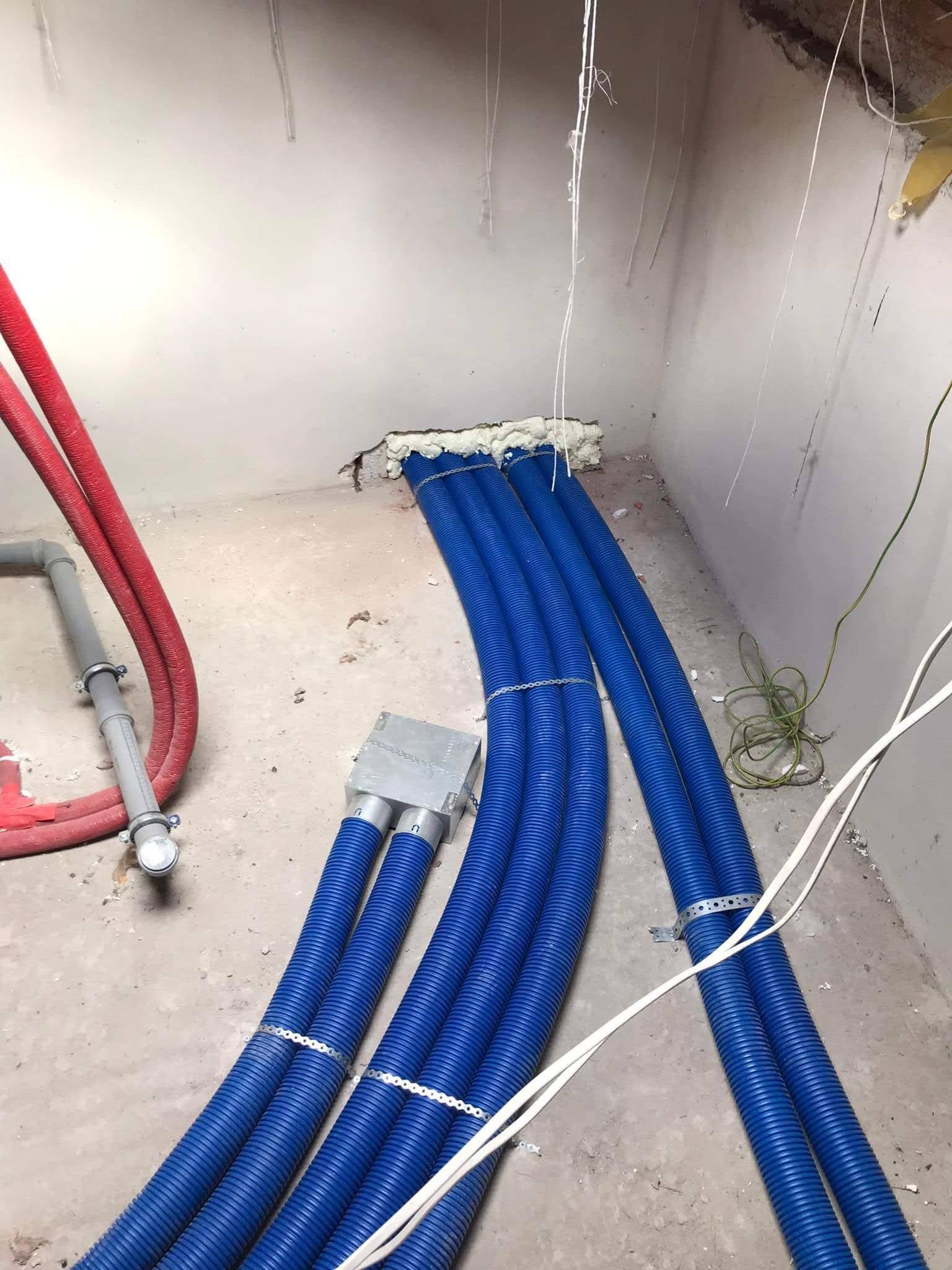 Wnętrze w trakcie prac instalacyjnych: wiązka niebieskich rur karbowanych przymocowanych metalowymi obejmami, wychodzących ze ściany uszczelnionej pianką montażową, obok czerwone rury i szare...