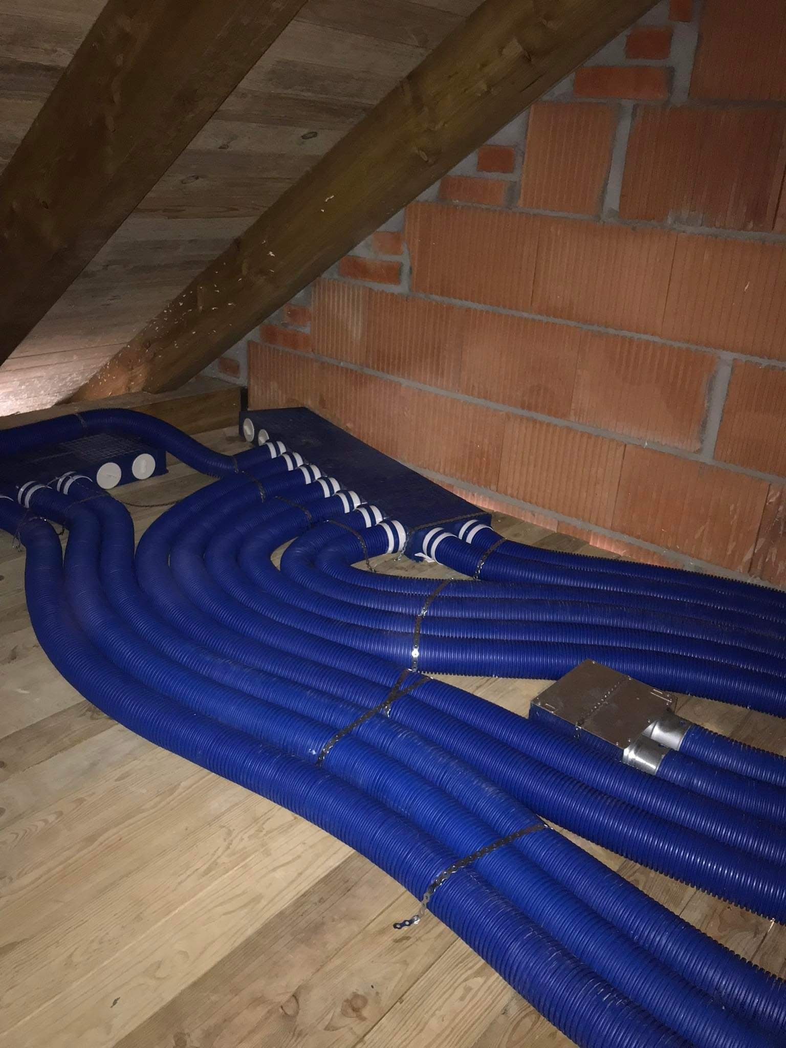Niebieskie, karbowane rury wentylacyjne ułożone równolegle na drewnianej podłodze poddasza, z widocznymi elementami mocującymi i skrzynką rozdzielczą.
