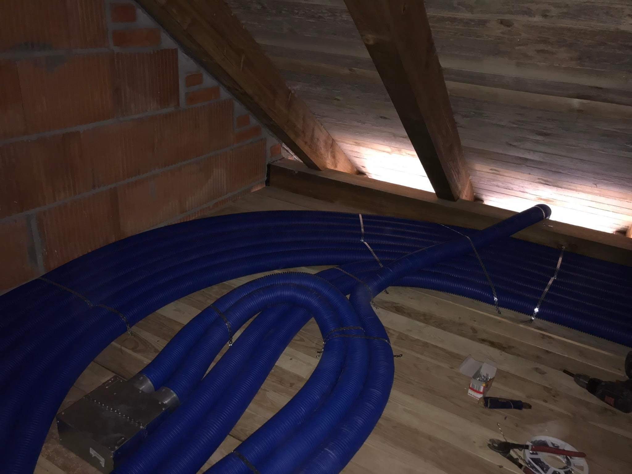 Niebieskie, karbowane przewody wentylacyjne rozprowadzone na drewnianej podłodze poddasza, z widoczną ceglaną ścianą i narzędziami montażowymi.