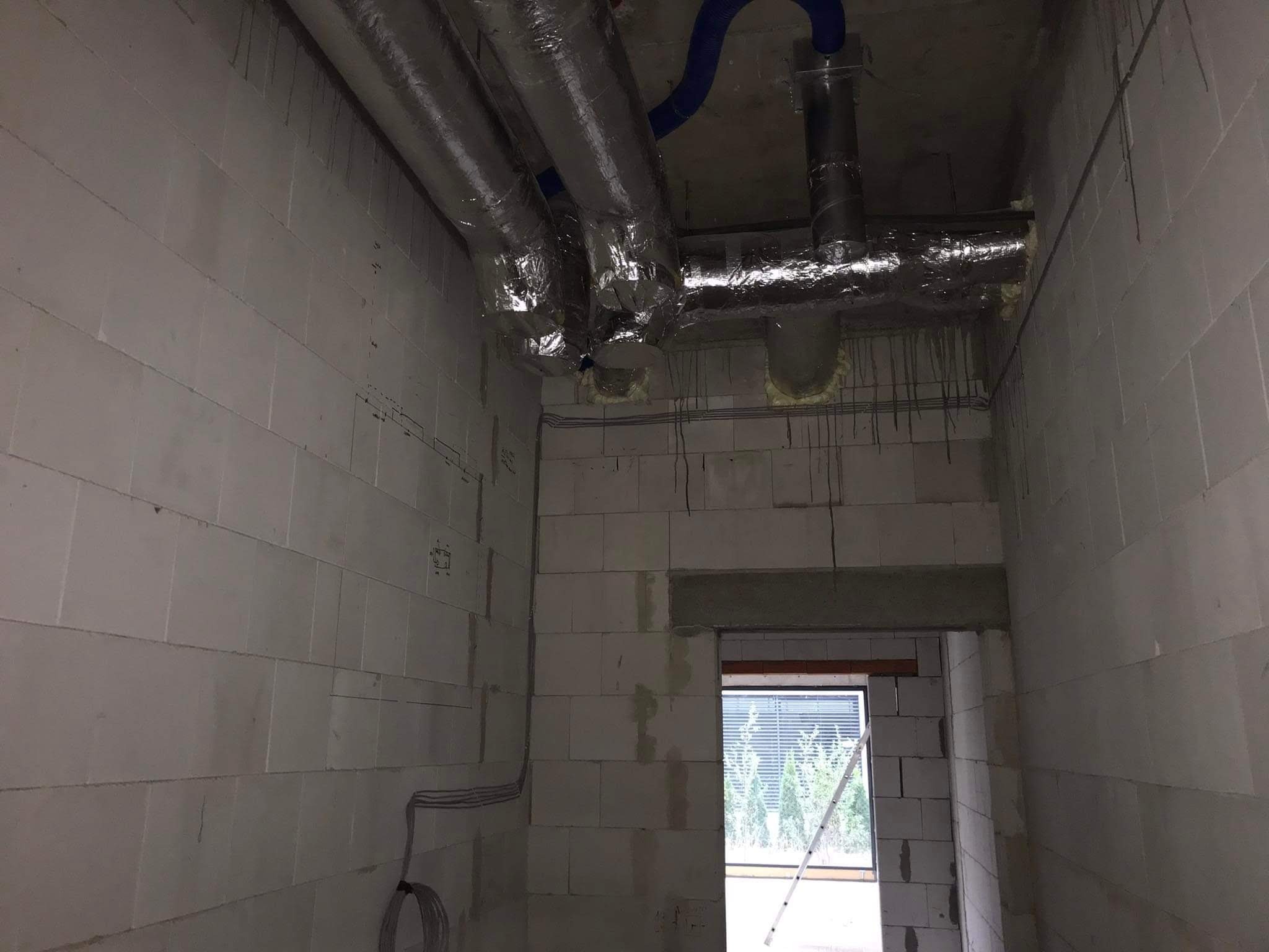Surowy stan budynku z widoczną instalacją wentylacyjną: srebrne rury izolowane folią aluminiową pod sufitem, niebieski elastyczny przewód wentylacyjny, ściany z białych bloczków, widok na zewnątrz...