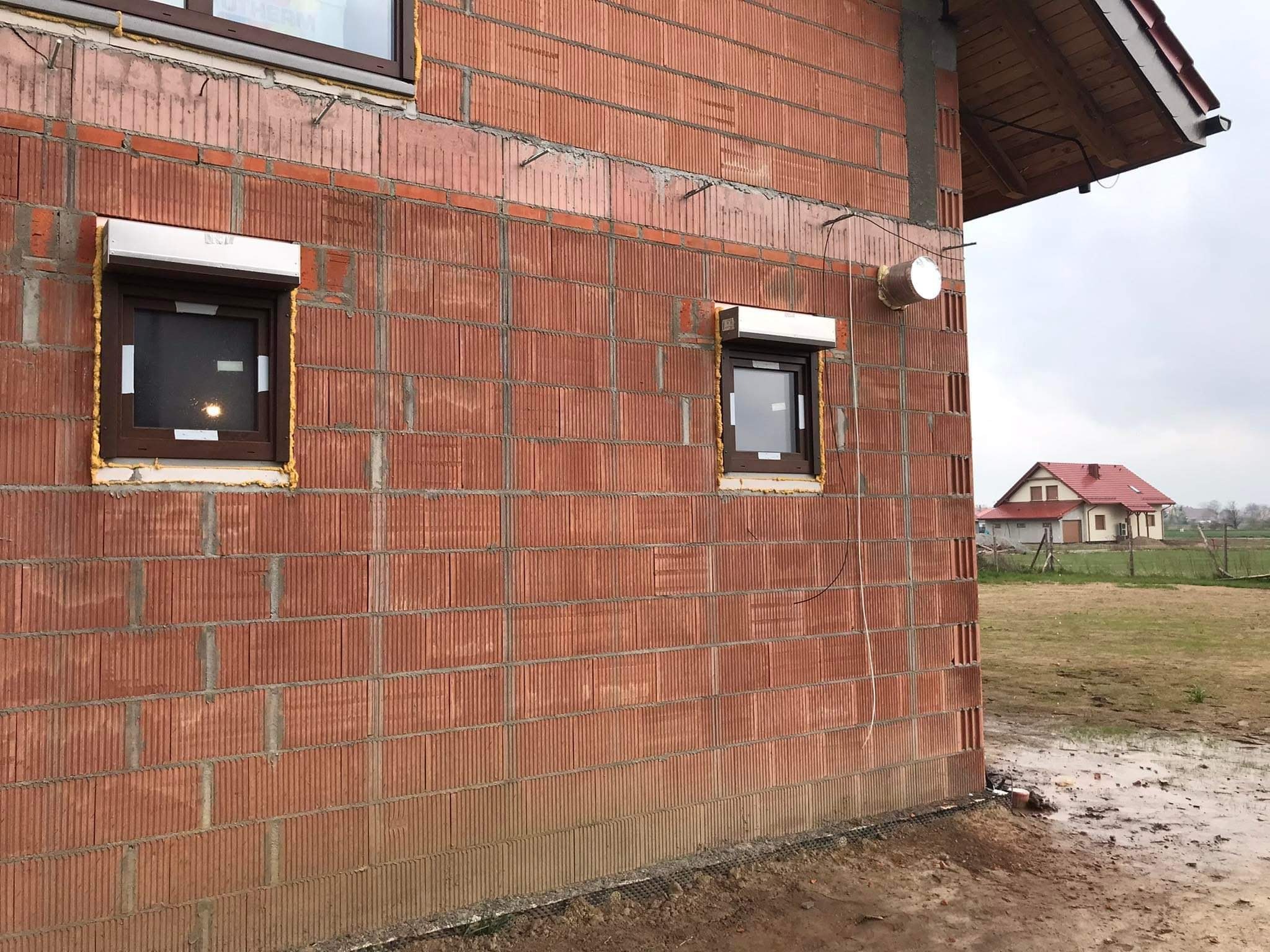 Surowy mur z czerwonej cegły ceramicznej z zamontowanymi oknami z brązowymi ramami i roletami zewnętrznymi, widoczne pianowanie uszczelniające, na zewnątrz budynku widoczny element instalacji...