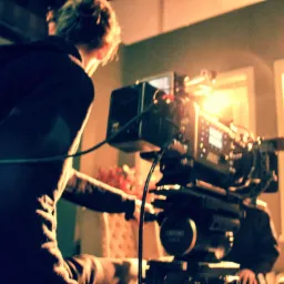 Operator filmowy obsługuje profesjonalną kamerę filmową z oświetleniem, w tle widoczne okna z żaluzjami.