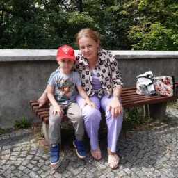 Chłopiec w czerwonej czapce i kobieta w kwiecistej bluzce siedzą obok siebie na drewnianej ławce na tle zieleni.