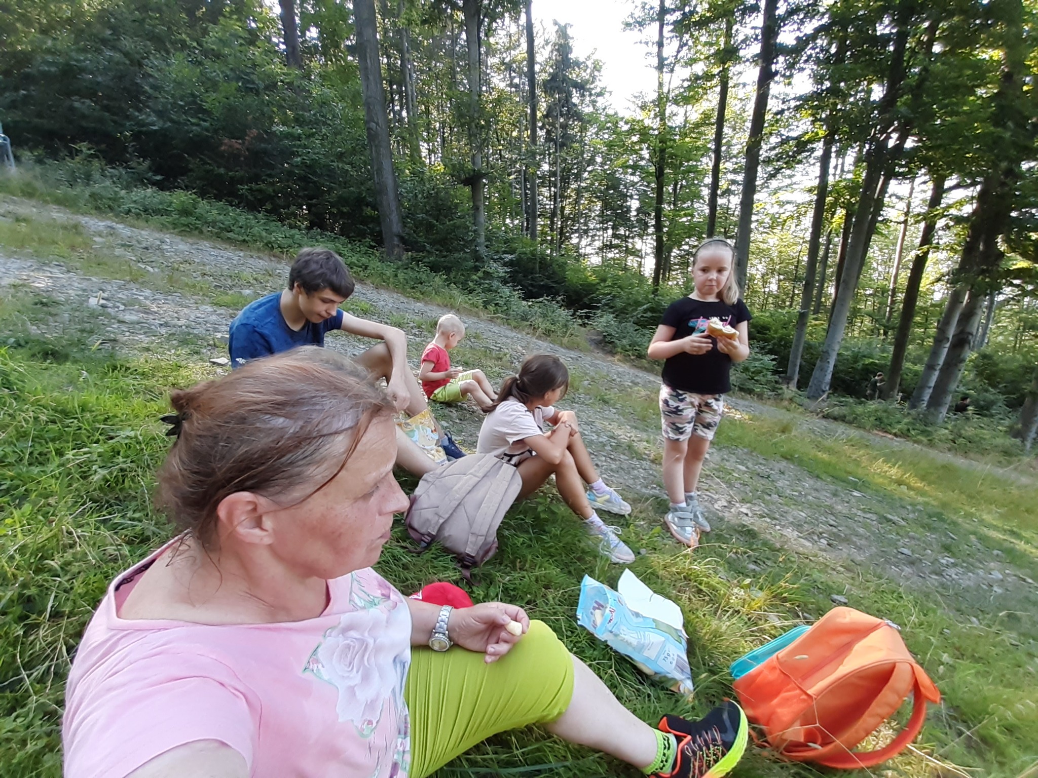 Grupa dzieci i dorosła kobieta siedzą na trawiastym zboczu w lesie, jedzą przekąski i odpoczywają.