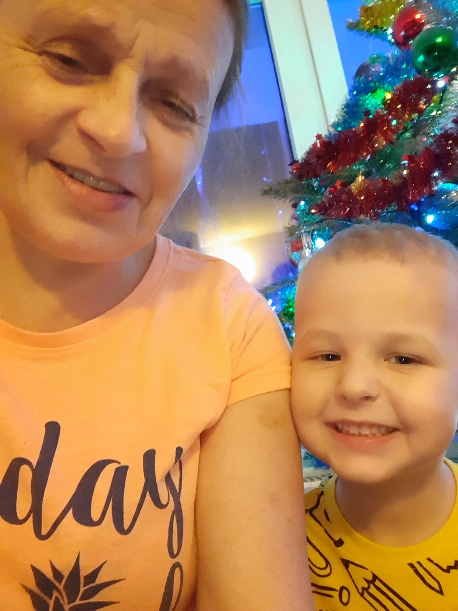 Uśmiechnięta kobieta w pomarańczowej koszulce z napisem 'day' i mały chłopiec w żółtej koszulce z nadrukiem ołówków, pozujący na tle choinki z kolorowymi światełkami.