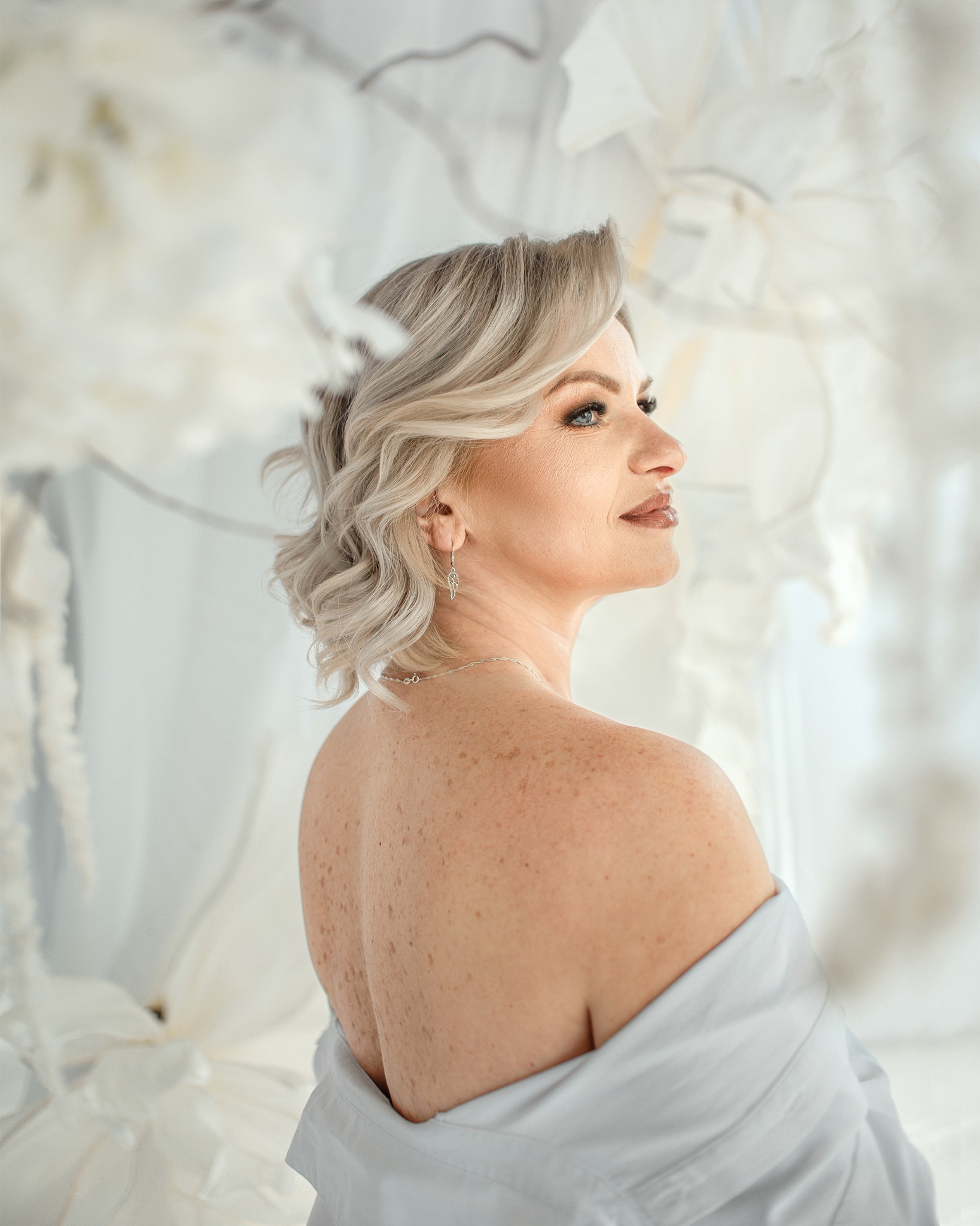 Portret kobiety z blond włosami, w białej koszuli, z widocznymi piegami na plecach, spoglądającej w bok na tle białych, dekoracyjnych kwiatów.