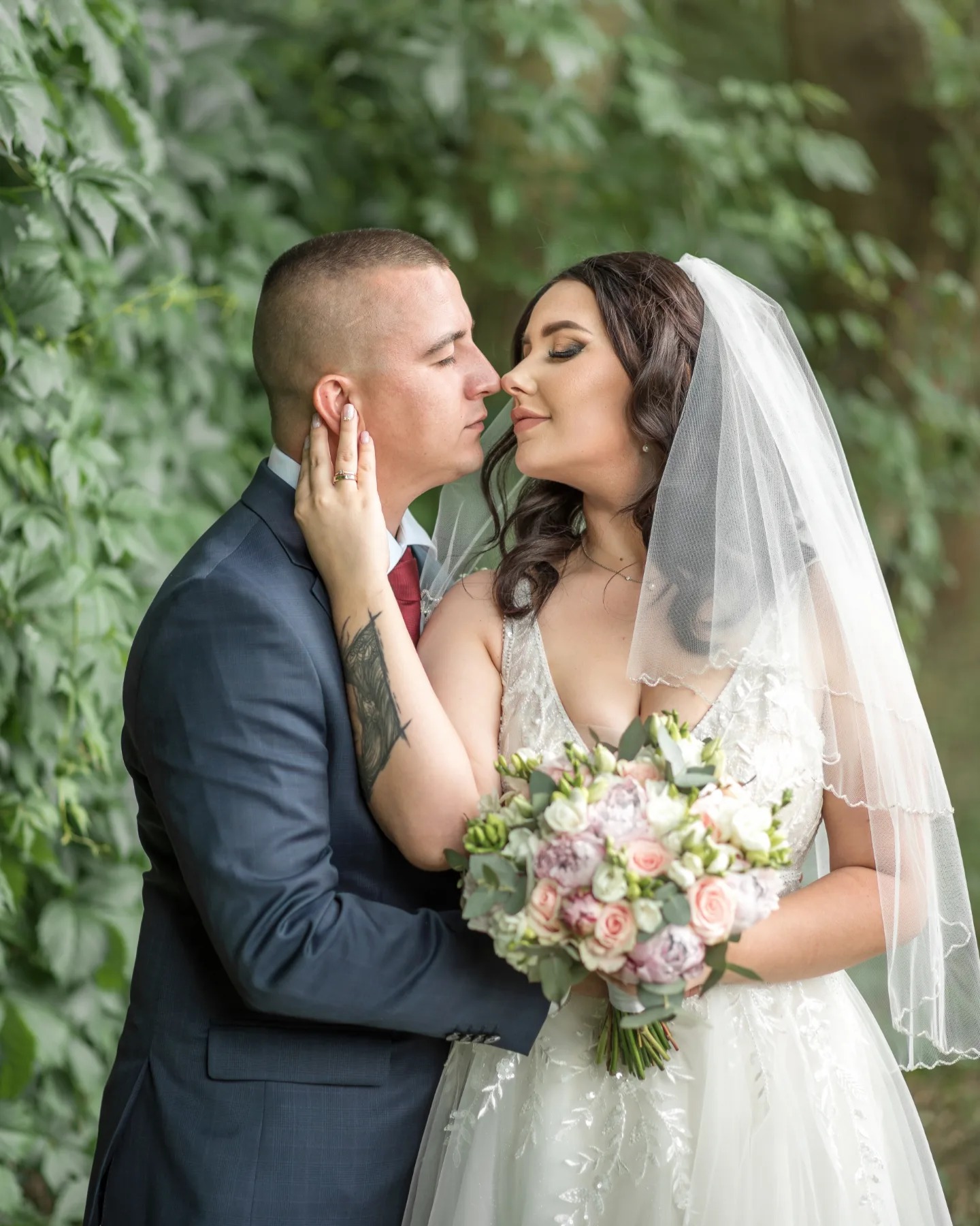 Para młoda delikatnie dotyka się nosami na tle zieleni, panna młoda trzyma bukiet różowych i białych kwiatów, widoczny tatuaż na ramieniu pana młodego.