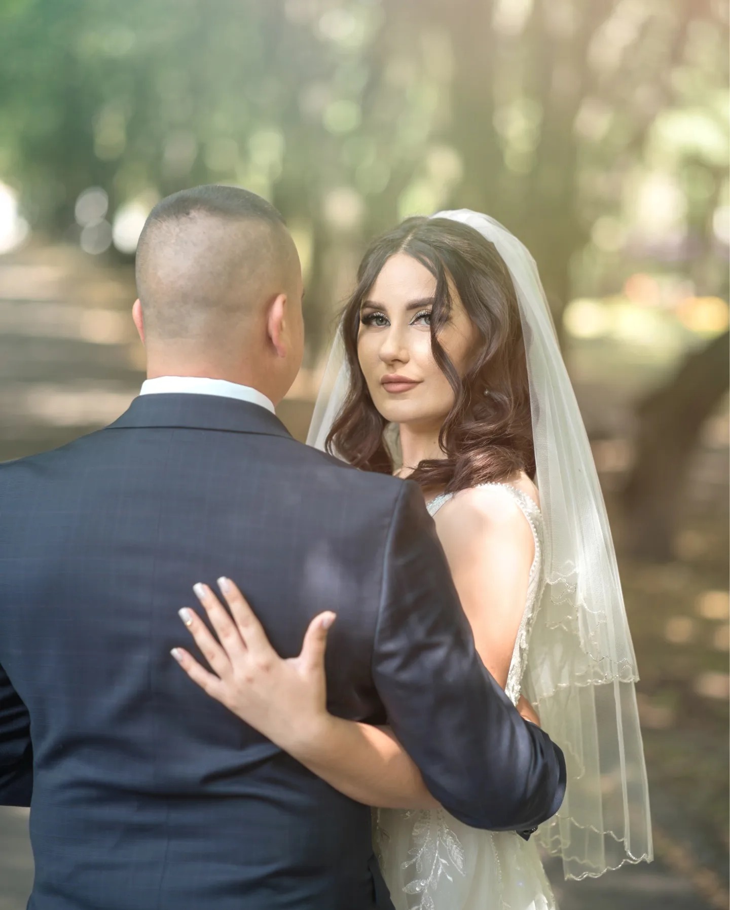 Panna młoda w białej sukni i welonie, obejmująca pana młodego w ciemnym garniturze, z delikatnym uśmiechem spogląda w obiektyw na tle rozmytej zieleni.