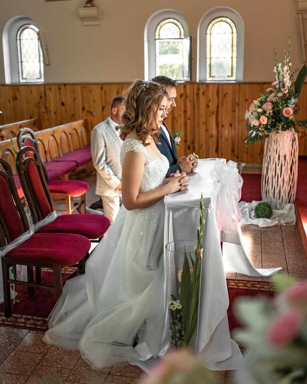 Para młoda w kościele podczas ceremonii ślubnej; panna młoda w białej sukni z tiulowym dołem i ozdobnym welonem, pan młody w ciemnym garniturze, oboje klęczą przed ołtarzem udekorowanym białym...