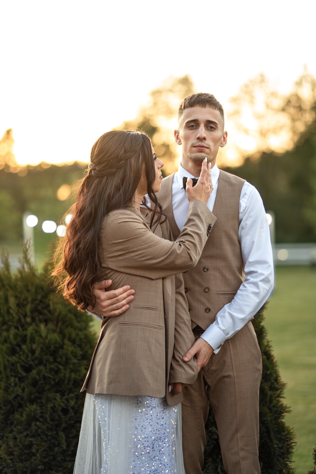 Elegancka para w dopasowanych brązowych strojach pozuje na tle zieleni i ciepłego światła zachodzącego słońca. Kobieta dotyka brody mężczyzny, tworząc intymną atmosferę.